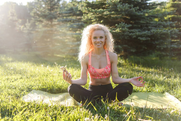 Image of young curly-haired sports woman practicing yoga on rug in park - Фото, изображение