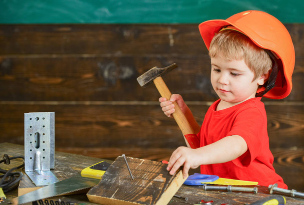 Toddler on busy face plays with hammer tool at home in workshop. Child in helmet cute playing as builder or repairer, repairing or handcrafting. Kid boy play as handyman. Handcrafting concept - Foto, imagen
