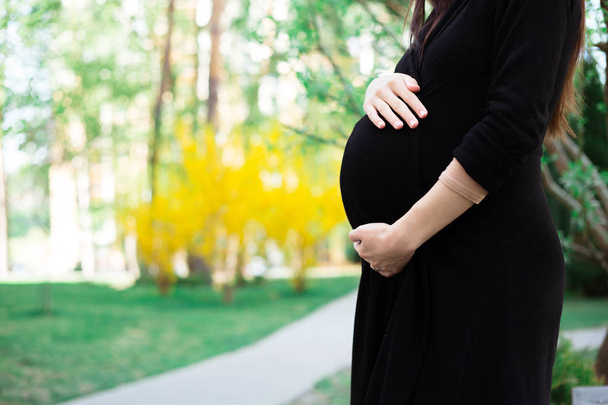 Mid section view of pregnant woman in black dress holding belly walking in park - Fotoğraf, Görsel