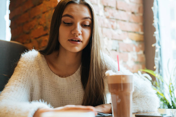 lovely long-haired girl freelancer in a white sweater sits in a cafe, works and drinks a fragrant cappuccino - Фото, зображення