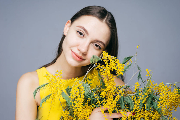 beautiful cute girl in yellow dress holds in hands a fragrant yellow mimosa, looks into camera and smiles - Fotografie, Obrázek