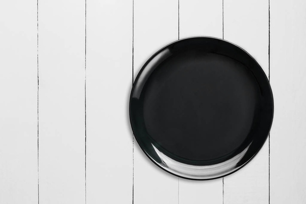 Placa de cerámica vacía negra sobre mesa de madera blanca, vista superior
. - Foto, Imagen