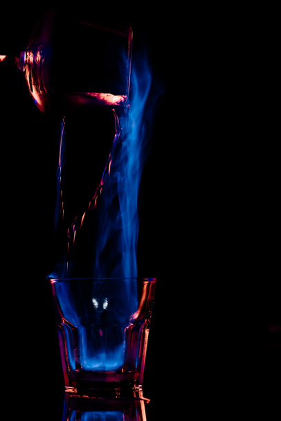 close up view of pouring burning sambuca alcohol drink into glass process on black background - Photo, Image