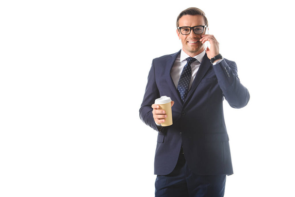 happy businessman in eyeglasses talking on smartphone and holding paper cup of coffee isolated on white background  - Foto, afbeelding