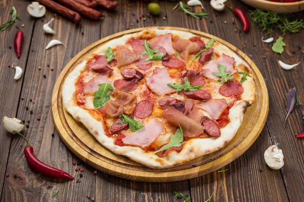 Delicious italian pizza with ham, bavarian sausages and prosciutto decorated by basil leaves on wooden background - 写真・画像