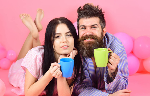 Man and woman in domestic clothes, pajamas. Man and woman on smiling faces lay, pink background. Couple in love drink coffee in bed. Couple relax in morning with coffee. Weekend morning concept - Foto, Imagen