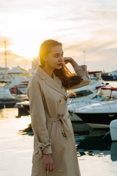розкішна молода жінка з довгим волоссям в модному бежевому пальто стоїть в морському порту ввечері і чекає свого корабля
 - Фото, зображення