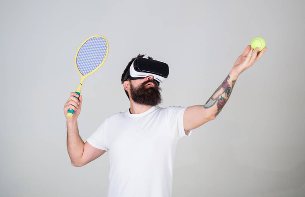 Handsome man in virtual reality headset playing tennis, grey background. Hipster use modern technology for sport games. Guy with VR glasses play tennis with racket and ball. Virtual tennis concept - Fotografie, Obrázek