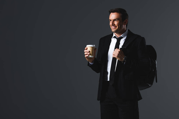 lachende zakenman met rugzak houden papieren kopje koffie geïsoleerd op een grijze achtergrond  - Foto, afbeelding
