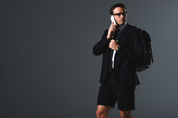 Selbstbewusster, stylischer Mann in kurzen Hosen mit Rucksack, der auf grauem Hintergrund mit dem Smartphone spricht  - Foto, Bild
