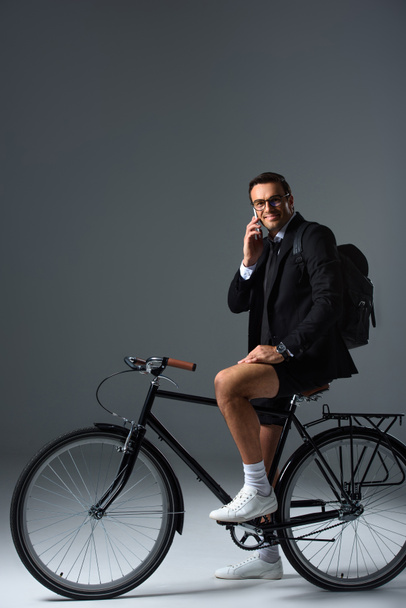 fashionable man with backpack sitting on bicycle talking on smartphone on grey background  - Zdjęcie, obraz