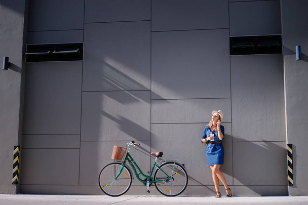 Young pretty woman using smartphone with bicycle leaning on the grey wall. - Foto, afbeelding