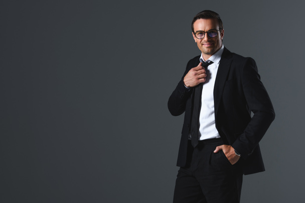 happy businessman in eyeglasses with hand in pocket adjusting tie isolated on grey background  - Fotografie, Obrázek