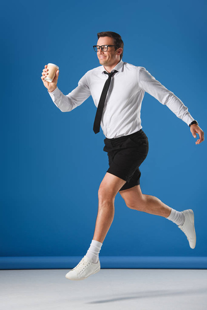 happy man holding disposable coffee cup while jumping and looking away on blue - Photo, image
