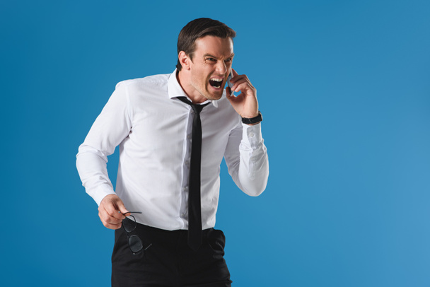 angry man holding eyeglasses and screaming while talking by smartphone isolated on blue - Photo, Image