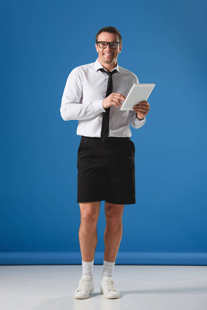 handsome businesman in eyeglasses and shorts using digital tablet and smiling at camera on blue - Foto, immagini