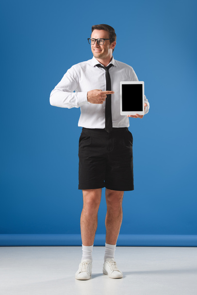 smiling man in eyeglasses and shorts pointing with finger at digital tablet with blank screen and looking away on blue - Fotó, kép