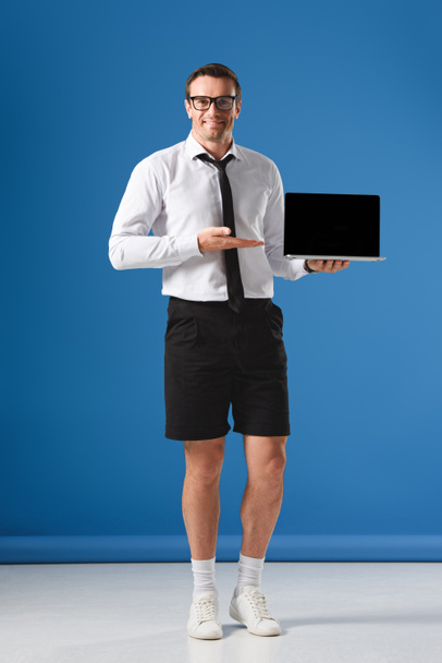 handsome businessman in shorts showing laptop with blank screen and smiling at camera on blue - Photo, Image