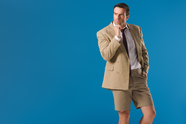 thoughtful man in shorts standing with hand on chin and looking away isolated on blue - Foto, Imagen