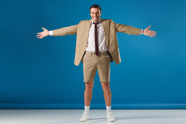 full length view of happy man in shorts standing with open arms and smiling at camera on blue - Foto, immagini