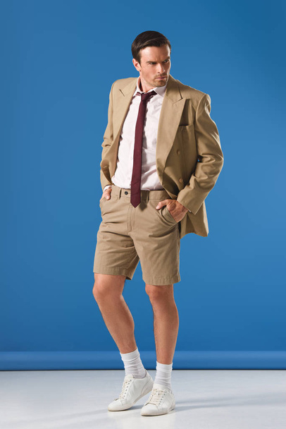 full length view of handsome man in suit jacket and shorts standing with hands in pockets and looking away on blue - Photo, Image