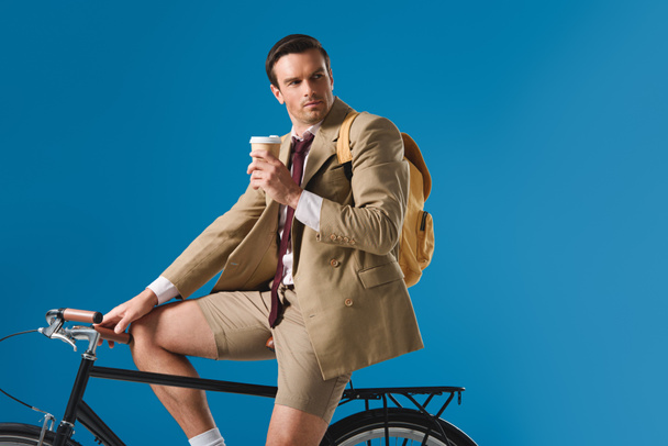 hombre sosteniendo taza de papel mientras monta bicicleta y mirando hacia otro lado aislado en azul
 - Foto, Imagen