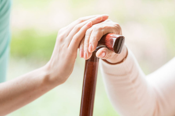 Close-up of caregiver holding hand of a senior person with walking stick against blurred background - Photo, Image