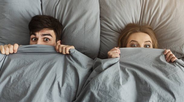 Σοκαρισμένος άνδρας και γυναίκα κρύβονται κάτω από την κουβέρτα στο κρεβάτι τους στο σπίτι - Φωτογραφία, εικόνα