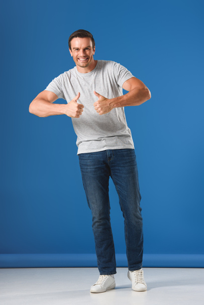 happy handsome man showing thumbs up and smiling at camera on blue - Foto, afbeelding