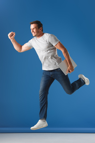hombre alegre sosteniendo el ordenador portátil y saltando sobre azul
 - Foto, Imagen