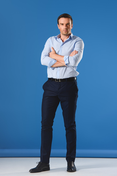 full length view of handsome man standing with crossed arms and looking at camera on blue - Foto, afbeelding