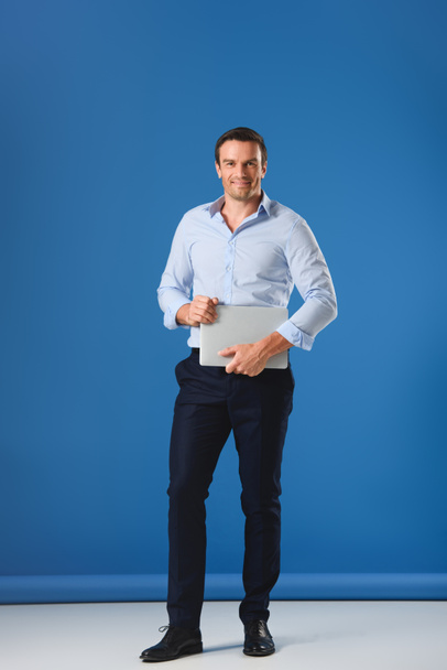 full length view of handsome man holding laptop and smiling at camera on blue - Fotoğraf, Görsel