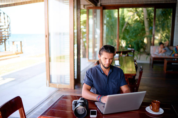 Young  bearded man working on laptop computer while sitting on cafe terrace. - 写真・画像
