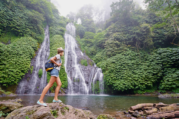 Young woman in hat with rucksack enjoying tropical waterfall view. - Foto, imagen