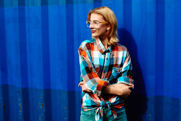 Портрет молодой блондинки в очках и клетчатой рубашке со скрещенными руками на фоне голубой железной стены
  - Фото, изображение