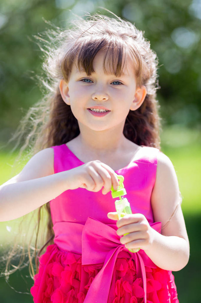 Beautiful girl in a red dress is playing with soap bubbles in a city park. A girl of 5 years old lets out soap bubbles. - Photo, Image