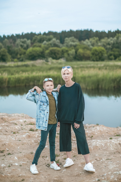 mooie stijlvolle moeder en zoon kijken camera terwijl je met een honkbalknuppel in de buurt van lake    - Foto, afbeelding