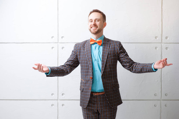 Cheerful young man in plaid suit and bowtie gesturing welcome sign and smiling while standing against white wall. - Photo, Image