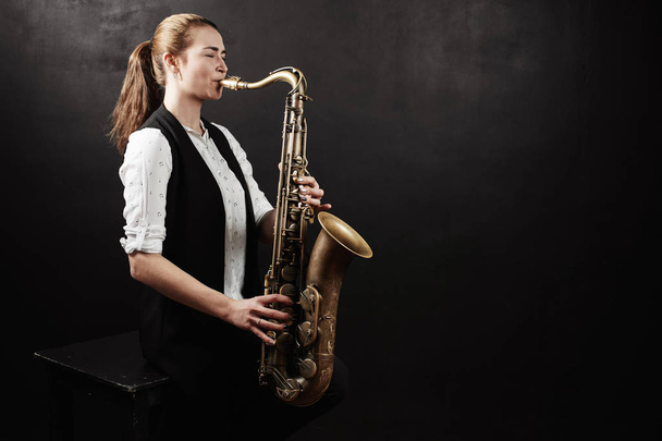 Young woman wearing white shirt and black vest playing saxophone on black background - Zdjęcie, obraz