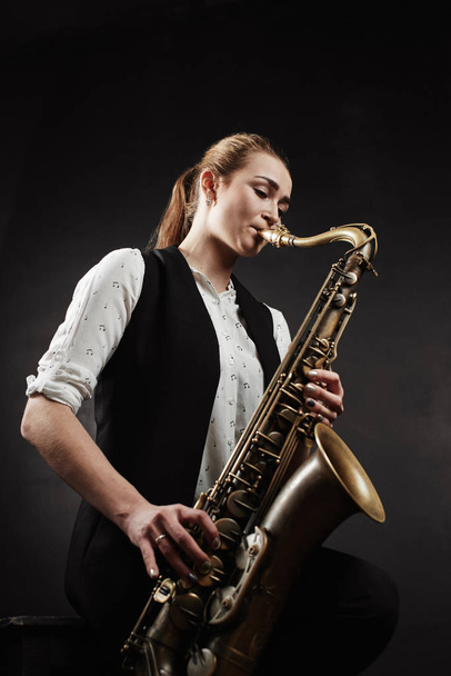 Young woman wearing white shirt and black vest playing saxophone on black background - Fotografie, Obrázek