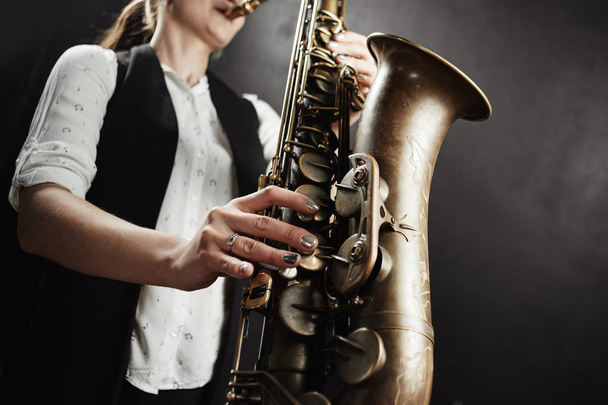 Young woman wearing white shirt and black vest playing saxophone on black background - Photo, Image
