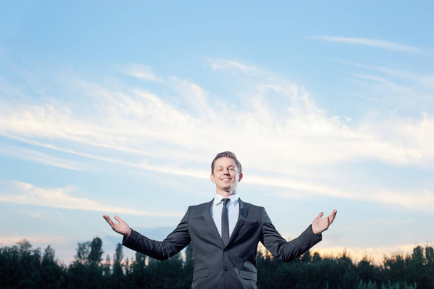 Happy confident young caucasian businessman in suit and tie keeping arms raised and expressing positivity while standing outdoors with sky in the background - 写真・画像