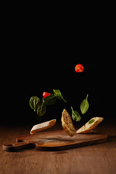 Bread pieces and salad leaves with tomatoes flying above wooden table surface - Photo, Image