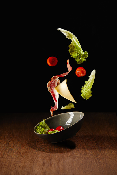 Fresh salad with meat and cheese falling in bowl above wooden table surface - Photo, Image