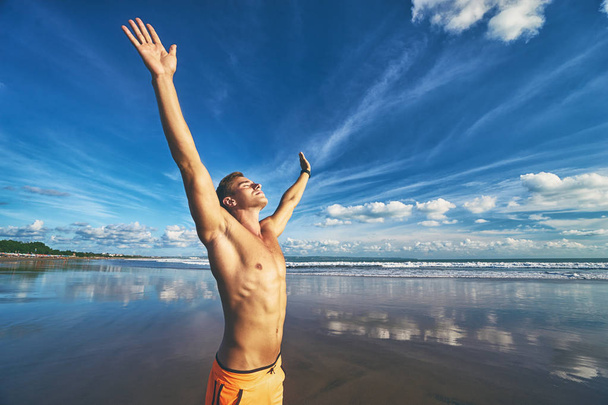 junger schöner starker Mann hebt die Hände am Strand gegen den Himmel. - Foto, Bild
