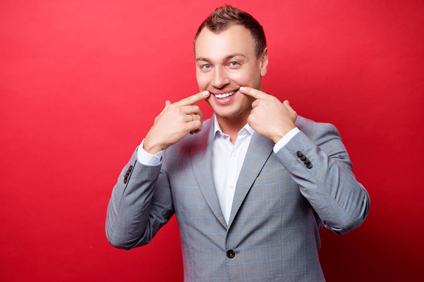 Handsome young business man keeping smile with his fingers while standing against red background. - Foto, imagen