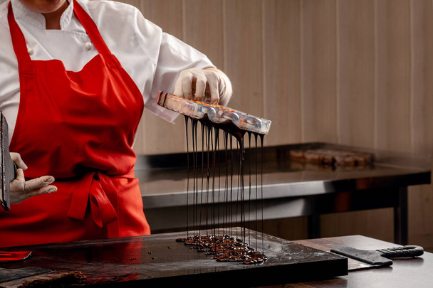 A woman confectioner with red uniform and white sterile gloves do a set of colorful chocolates from milk chocolate on a table. - Foto, Imagem