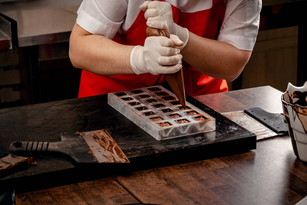 Bir kadın pudra kırmızı üniforma ve beyaz steril eldiven ile sütlü çikolata bir tablo üzerinden bir dizi renkli çikolata yapmak. - Fotoğraf, Görsel