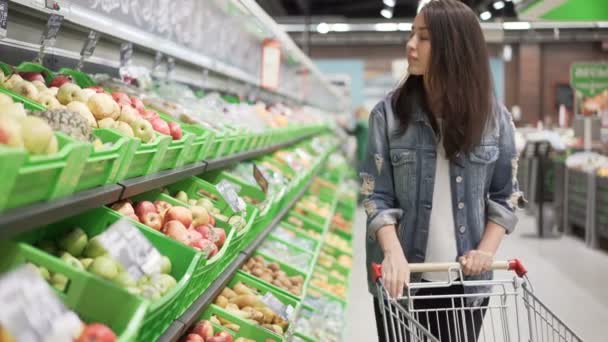 Pretty young woman is choosing fruit in grocery store, she is touching and smelling apples then putting them in trolley. Healthy food, beautiful girls and shops concept. - Video