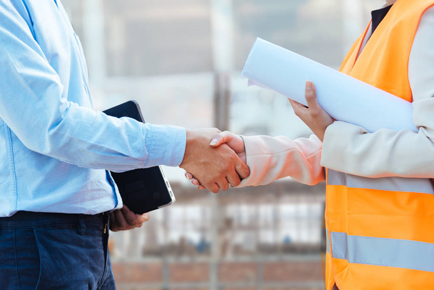Close up Asian man civil engineer holding laptop and Asian woman architect holding drawing blueprint shaking hands at construction site. - Foto, imagen